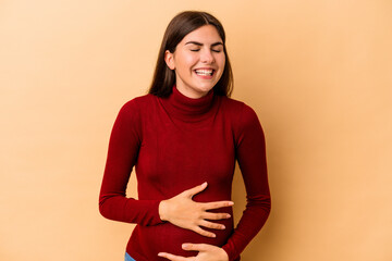Obraz premium Young caucasian pregnant woman isolated on beige background laughs happily and has fun keeping hands on stomach.