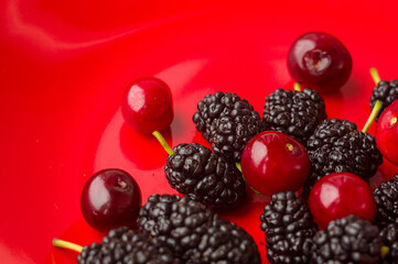 Ripe large black mulberries and red cherries on a red plate, close-up.