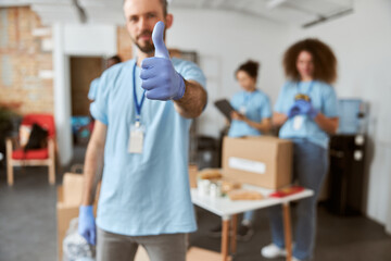 Male volunteer in blue uniform and protective gloves showing thumbs up at camera. Team sorting, packing items in cardboard boxes