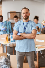 Fototapeta premium Confident caucasian male volunteer in blue uniform and protective gloves standing with arms crossed. Team sorting, packing items in cardboard boxes