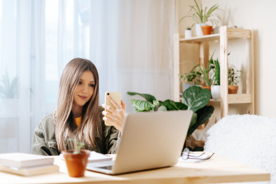 Freelance woman with mobile cell phone typing at laptop and working from home office with plants. Happy girl on workplace at the desk taking selfie. Distance learning online education and work.