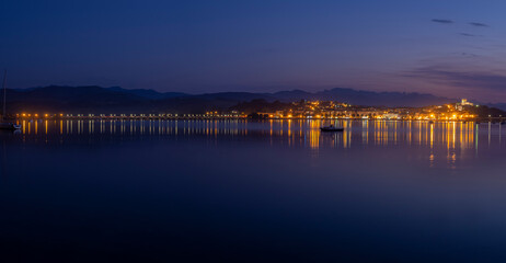Fototapeta na wymiar Panorámica nocturna del puente de San Vicente de la Barquera iluminado , con los reflejos de las luces de colores en el mar, y las barcas meciéndose en el agua, en Santander, España, verano de 2020.
