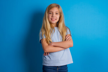 Fototapeta na wymiar beautiful caucasian little girl wearing blue T-shirt over blue background being happy smiling and crossed arms looking confident at the camera. Positive and confident person.