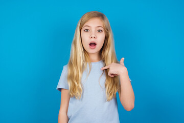 beautiful Caucasian little girl wearing white T-shirt over blue background being in stupor shocked, has astonished expression pointing at oneself with finger saying: Who me?