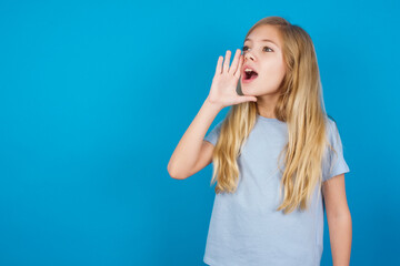 beautiful Caucasian little girl wearing blue T-shirt over blue background profile view, looking happy and excited, shouting and calling to copy space.