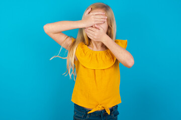 Caucasian kid girl wearing yellow T-shirt against blue wall Covering eyes and mouth with hands, surprised and shocked. Hiding emotions.