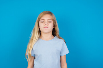 beautiful Caucasian little girl wearing blue T-shirt over blue background crosses eyes, puts lips, makes grimace with awkward expression has fun alone, plays fool.