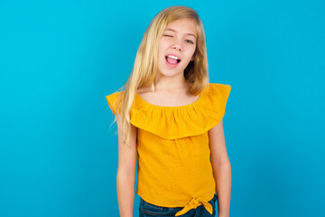Caucasian kid girl wearing yellow T-shirt against blue wall winking looking at the camera with sexy...