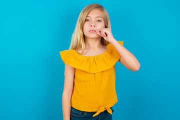 Caucasian kid girl wearing yellow T-shirt against blue wall mouth and lips shut as zip with fingers. Secret and silent, taboo talking. © Roquillo