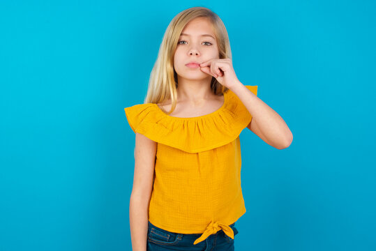 Caucasian kid girl wearing yellow T-shirt against blue wall mouth and lips shut as zip with fingers. Secret and silent, taboo talking.