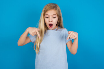 Amazed beautiful Caucasian little girl wearing blue T-shirt over blue background points down with...