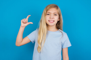 Fototapeta premium beautiful Caucasian little girl wearing white T-shirt over blue background smiling and gesturing with hand small size, measure symbol.