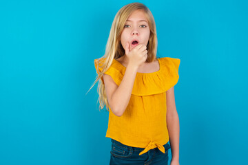 Caucasian kid girl wearing yellow T-shirt against blue wall Looking fascinated with disbelief, surprise and amazed expression with hands on chin