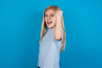 Overjoyed beautiful Caucasian little girl wearing blue T-shirt over blue background glad to receive good news, clenching fist and making winning gesture.