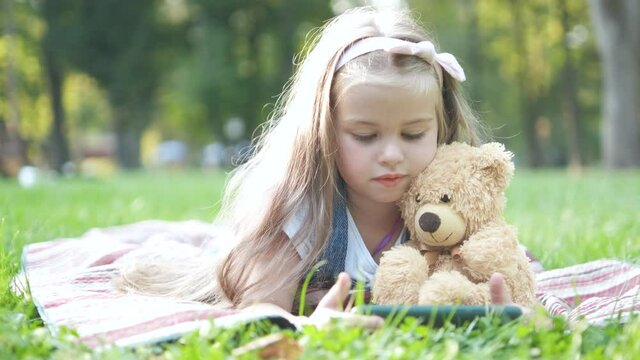 Small kid girl looking in her mobile phone together with her favorite teddy bear toy outdoors in summer park.