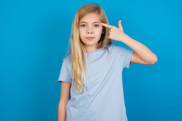 Fototapeta premium Unhappy beautiful Caucasian little girl wearing blue T-shirt over blue background curves lips and makes suicide gesture, shoots in temple with hand, tries kill himself.