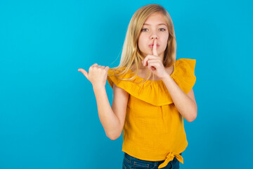 Obraz premium Caucasian kid girl wearing yellow T-shirt against blue wall asking to be quiet with finger on lips pointing with hand to the side. Silence and secret concept.