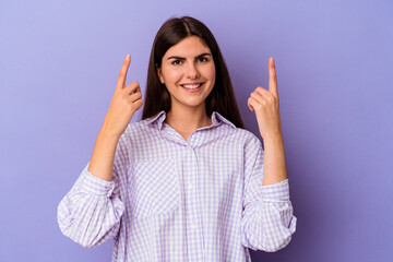 Young caucasian woman isolated on purple background indicates with both fore fingers up showing a blank space.