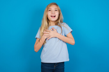 Fototapeta premium Happy smiling beautiful Caucasian little girl wearing blue T-shirt over blue background has hands on chest near heart. Human emotions, real feelings and facial expression concept.