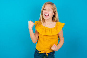 Caucasian kid girl wearing yellow T-shirt against blue wall very happy and excited doing winner gesture with arms raised, smiling and screaming for success. Celebration concept.