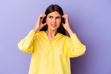 Young caucasian woman isolated on purple background covering ears with fingers, stressed and desperate by a loudly ambient.