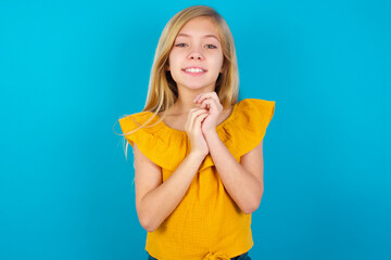 Obraz premium Positive Caucasian kid girl wearing yellow T-shirt against blue wall smiles happily, glad to receive pleasant news from interlocutor, keeps hands together. People emotions concept.