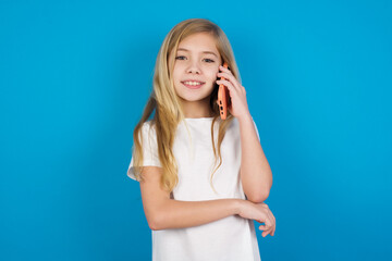 Fototapeta premium Portrait of a smiling beautiful Caucasian little girl wearing white T-shirt over blue background talking on mobile phone. Business, confidence and communication concept.