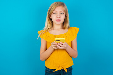 Caucasian kid girl wearing yellow T-shirt against blue wall enjoys distant communication, uses mobile phone, surfs fast unlimited internet, has pleasant smile, makes shopping online,