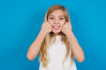 Fototapeta premium Happy beautiful Caucasian little girl wearing white T-shirt over blue background keeps fists on cheeks smiles broadly and has positive expression being in good mood