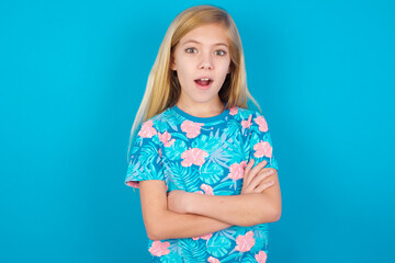 Shocked embarrassed Caucasian kid girl wearing hawaiian T-shirt against blue wall keeps mouth widely opened. Hears unbelievable novelty stares in stupor