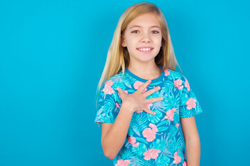 Caucasian kid girl wearing hawaiian T-shirt against blue wall smiles toothily cannot believe eyes expresses good emotions and surprisement