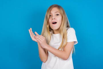 Surprised emotional beautiful Caucasian little girl wearing white T-shirt over blue background rubs palms and stares at camera with disbelief