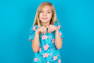 Caucasian kid girl wearing hawaiian T-shirt against blue wall makes bunny paws and looks with...