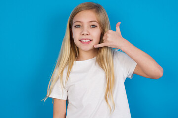 beautiful Caucasian little girl wearing white T-shirt over blue background makes phone gesture, says call me back again, has glad expression.
