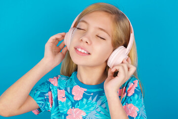 Pleased Caucasian kid girl wearing hawaiian T-shirt against blue wall enjoys listening pleasant melody keeps hands on stereo headphones closes eyes. Spending free time with music