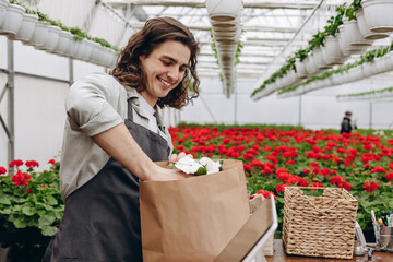 Portrait of male florist selling flowerpot and putting it in eco bag in garden centre. Flower shop.