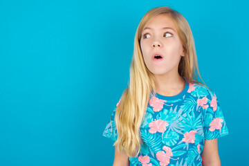 Shocked Caucasian kid girl wearing hawaiian T-shirt against blue wall look empty space with open mouth screaming: Oh My God! I can't believe this.