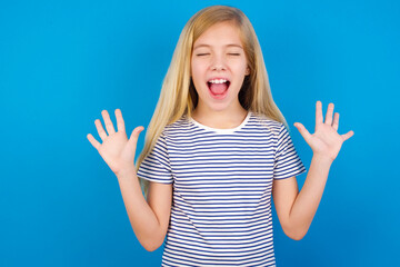 Fototapeta premium Emotive Caucasian kid girl wearing striped shirt ​against blue wall laughs loudly, hears funny joke or story, raises palms with satisfaction, being overjoyed amused by friend