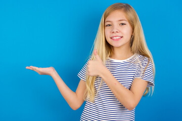 Fototapeta premium Caucasian kid girl wearing striped shirt ​against blue wall Showing palm hand and doing ok gesture with thumbs up, smiling happy and cheerful.