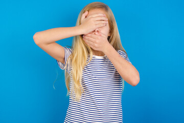 Caucasian kid girl wearing striped shirt ​against blue wall Covering eyes and mouth with hands, surprised and shocked. Hiding emotions.