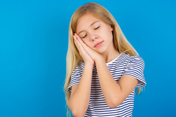 Caucasian kid girl wearing striped shirt ​against blue wall  sleeping tired dreaming and posing with hands together while smiling with closed eyes.