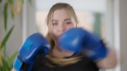 Young overweight woman looking at camera boxing shadow. Portrait of concentrated focused Caucasian female boxer training at home indoors. Hobby and sport lifestyle