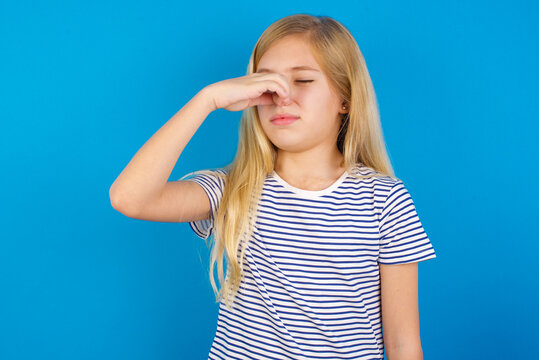 Caucasian kid girl wearing striped shirt ​against blue wall  smelling something stinky and disgusting, intolerable smell, holding breath with fingers on nose. Bad smell