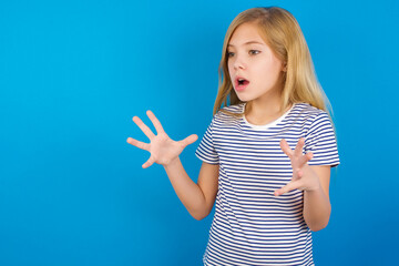 Fototapeta premium Caucasian kid girl wearing striped shirt ​against blue wall shouts loud, keeps eyes opened and hands tense.