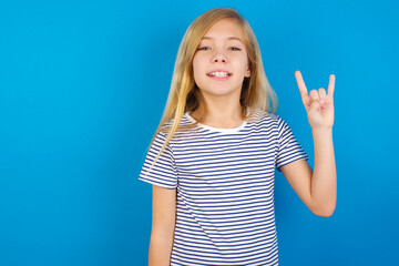 Caucasian kid girl wearing striped shirt ​against blue wall  doing a rock gesture and smiling to the camera. Ready to go to her favorite band concert.