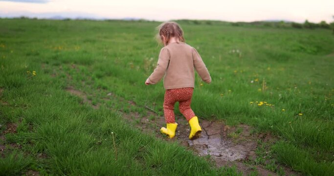 cute toddler girl having fun in blooming spring field, playing in muddy puddle