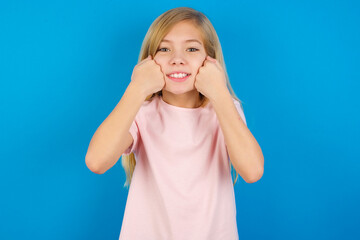 Happy Caucasian kid girl wearing pink shirt against blue wall keeps fists on cheeks smiles broadly and has positive expression being in good mood