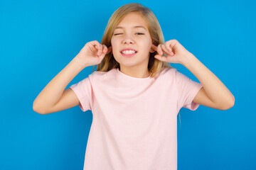 Happy Caucasian kid girl wearing pink shirt against blue wall ignores loud music and plugs ears with fingers asks to turn off sound