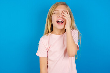 Caucasian kid girl wearing pink shirt against blue wall makes face palm and smiles broadly, giggles positively hears funny joke poses