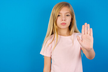 Caucasian kid girl wearing pink shirt against blue wall shows stop sign prohibition symbol keeps...